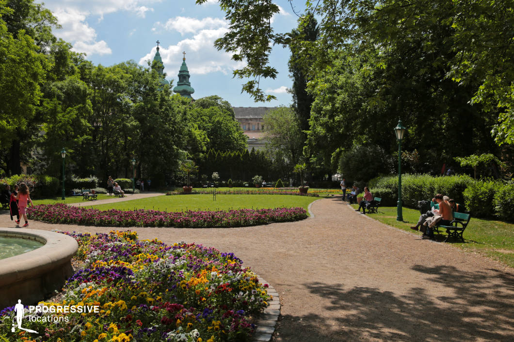 nal blooms, neatly trimmed hedges, and a manicured lawn. A winding gravel path lined with classic green lampposts and wooden benches leads through the lush greenery, offering a serene and structured aesthetic. The setting is framed by mature trees providing natural shade, while in the background, historic architecture with distinctive spires and ornate facades rises above the foliage, adding a sense of timeless elegance. This location is ideal for refined outdoor shoots, period-style productions, or tranquil promotional visuals highlighting classical and historic urban parks.