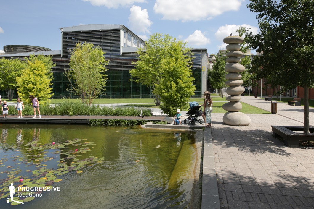 The image showcases a vibrant urban park with a calm water feature reflecting the surrounding lush greenery. The focus is on the beautiful integration of nature and modern architectural elements. A serene pond, with delicate lily pads floating on its surface, mirrors the surrounding trees and the nearby modern glass building, creating a harmonious balance between natural and contemporary design. The park features a sleek, well-maintained wooden walkway extending into the water, surrounded by tall, leafy trees with bright green foliage. The trees provide a sense of shelter and comfort, with their rich green canopies adding a feeling of freshness and vibrancy to the space. Alongside the pathway, decorative plantings and a towering stone sculpture offer an artistic contrast to the natural environment, emphasizing the blending of organic and man-made elements. The photograph captures a peaceful atmosphere, perfect for relaxation and contemplation. The clear water, lush plants, and the contemporary sculpture evoke a sense of calm and modernity, making this an ideal location for peaceful outdoor activities, reflecting the seamless coexistence of nature and urban spaces.