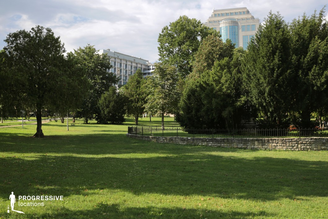 This image depicts a spacious park with vibrant greenery, framed by large trees that provide shade and add structure to the scene. The expansive grassy area creates a serene atmosphere, with a stone fence in the foreground adding an architectural touch. In the background, a collection of modern buildings with glass facades creates a striking contrast with the natural environment, subtly emphasizing the park’s integration within an urban landscape. The partly cloudy sky bathes the park in soft light, enhancing the peaceful ambiance. This park’s blend of lush vegetation and contemporary architecture offers a versatile setting for various film scenes, ideal for capturing moments of tranquility amid the surrounding cityscape.