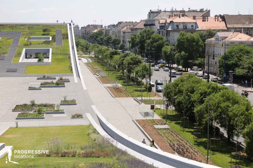  This photograph captures a modern urban park with expansive green spaces, offering a striking contrast between nature and architecture. The sloping roof of the park's central structure seamlessly blends into the green landscape, providing a unique visual experience. The clean, geometric lines of the pathways and raised garden beds contrast with the surrounding trees and urban backdrop. The busy street in the distance, lined with trees, adds a dynamic urban context while maintaining the serenity of the park's open space. The park is an ideal location for a film scene, as it embodies a modern and calm atmosphere, suitable for both intimate shots and broader cityscapes. The combination of nature and sleek modern design makes this spot versatile for many creative projects.