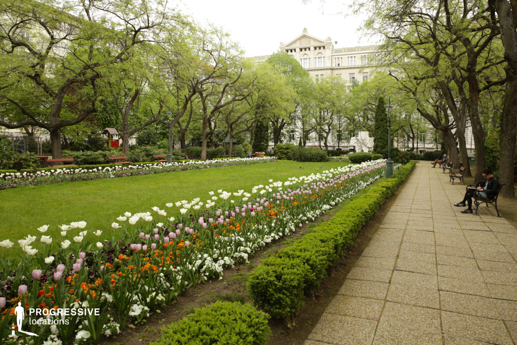  This city park scene features well-maintained flower beds lined with white, pink, and orange tulips, providing a vibrant contrast to the manicured green lawns. The path, paved with gravel, runs through the park and is bordered by neatly trimmed hedges. Rows of trees, still in the early stages of blooming, frame the space and add a natural canopy, enhancing the peaceful atmosphere. A few park benches are placed along the path, creating perfect spots for quiet reflection or relaxation. The background reveals a classic, elegant building, emphasizing the park's connection to the urban environment. This location is ideal for serene or introspective film scenes, with its balance of greenery and architectural elements. The spacious, symmetrical layout offers versatility for a variety of shots, from wide landscape views to more intimate close-ups along the benches. The park's calm, orderly environment makes it suitable for a range of creative projects, from romantic dramas to reflective moments in urban settings.