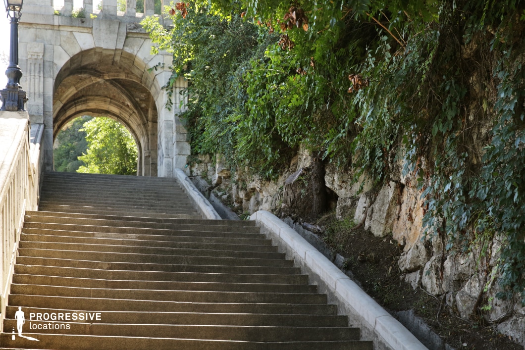 This image captures a set of stone stairs leading up beneath a grand archway, flanked by lush greenery and rugged rock formations. The smooth, symmetrical stairs create a sense of movement and depth, drawing the eye upward toward the arch, which acts as a natural frame for the view beyond. On either side, dense ivy and foliage grow along the stone walls, adding a touch of nature’s organic beauty to the architectural structure. The stairs and archway create a compelling visual contrast between the man-made and natural elements, providing a versatile backdrop for cinematic moments. The layout and perspective lend themselves to dramatic, upward shots, ideal for action sequences or scenes focused on ascension or progression. This location could be used for a variety of film genres, including suspenseful or reflective scenes, due to its layered, immersive structure. The combination of architectural elegance and natural elements offers a visually rich environment perfect for dynamic storytelling.