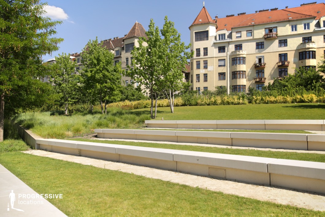 This contemporary park features a clean, geometric layout with a series of stepped concrete benches, offering a minimalistic yet functional design. The benches are arranged in parallel rows, creating a dynamic pattern against the lush green grass, with the topography gradually rising in the background. A line of young trees provides a subtle, natural contrast to the urban environment. The residential buildings in the background, with their traditional architecture and sloped roofs, add a sense of history and character to the setting. The park’s smooth, open space and modern features make it a versatile location for a variety of cinematic purposes, from urban drama to minimalist storytelling. The combination of the sculptural benches, greenery, and classical architecture provides a visual balance that can be used for both wide shots and more focused close-ups. The layout offers multiple possibilities for movement and interaction, ideal for scenes requiring subtle interaction with the environment or urban exploration.