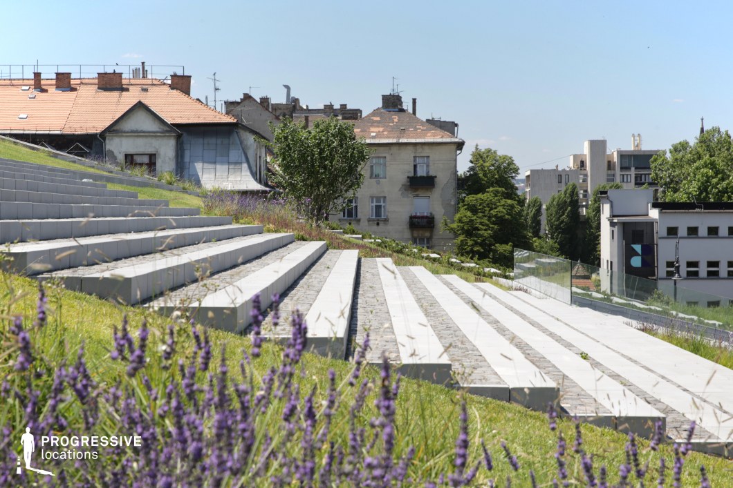 This contemporary urban scene features a set of wide, stepped terraces, leading up a gently sloping hill. The stairs are constructed with large, clean stone blocks interspersed with cobblestones, creating a modern yet rustic look. The surrounding greenery, including lavender plants, adds a touch of color and texture to the scene, complementing the minimalist design of the stairs. In the background, a variety of modern buildings, with both traditional and sleek architectural styles, rise above the park, creating a dynamic urban environment. This location provides an excellent setting for scenes that require an elegant, modern urban atmosphere. The wide, open stairs offer a versatile space for both wide-angle shots capturing the surrounding architecture and nature, as well as close-ups highlighting the interplay between the steps, the plants, and the cityscape beyond. The combination of modern design elements with natural surroundings makes it suitable for a variety of cinematic styles, from contemporary dramas to architectural features.