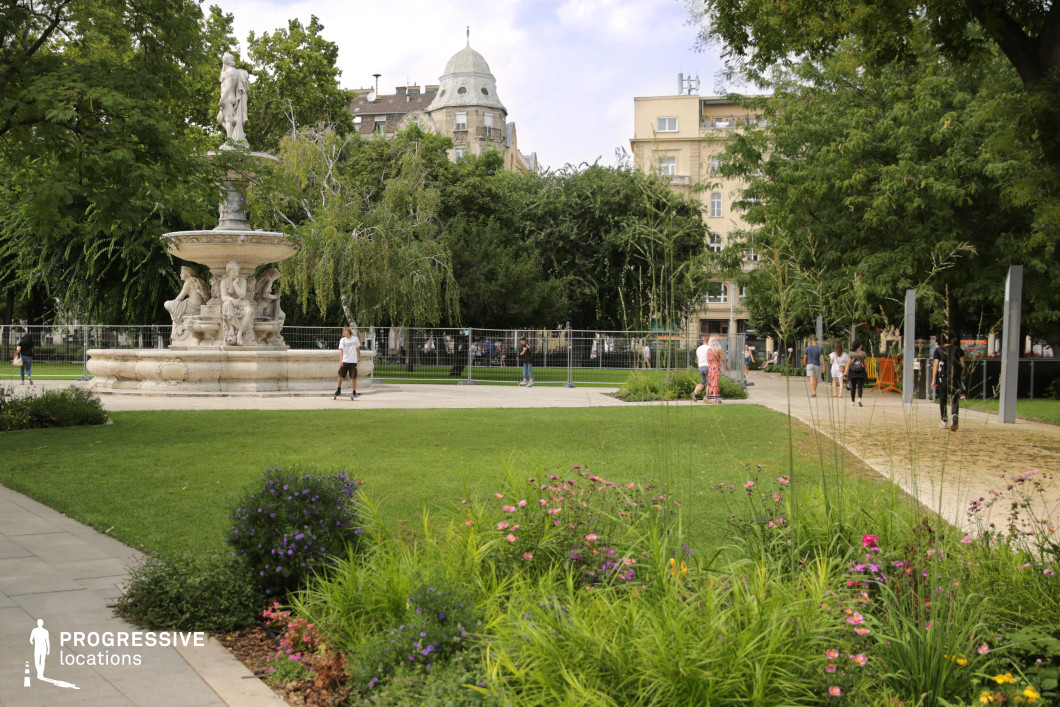 This elegant park scene features a large, classical stone fountain with intricate sculptural details, surrounded by lush greenery and vibrant flowers. The fountain, with its beautifully carved figures, stands as a central visual focal point, surrounded by well-maintained grass and colorful floral beds that enhance the natural beauty of the space. In the background, a mix of trees and modern buildings creates a balanced urban environment, with the tall trees offering shade and texture to the scene. This location provides a versatile setting, ideal for cinematic shots that require a classical architectural element combined with nature. The fountain’s detailed sculptures and the surrounding garden make this an excellent location for visually rich wide shots or focused close-ups of the fountain and its details. The overall serene ambiance of the park, with its combination of traditional and natural beauty, offers a peaceful setting suitable for a variety of creative projects.