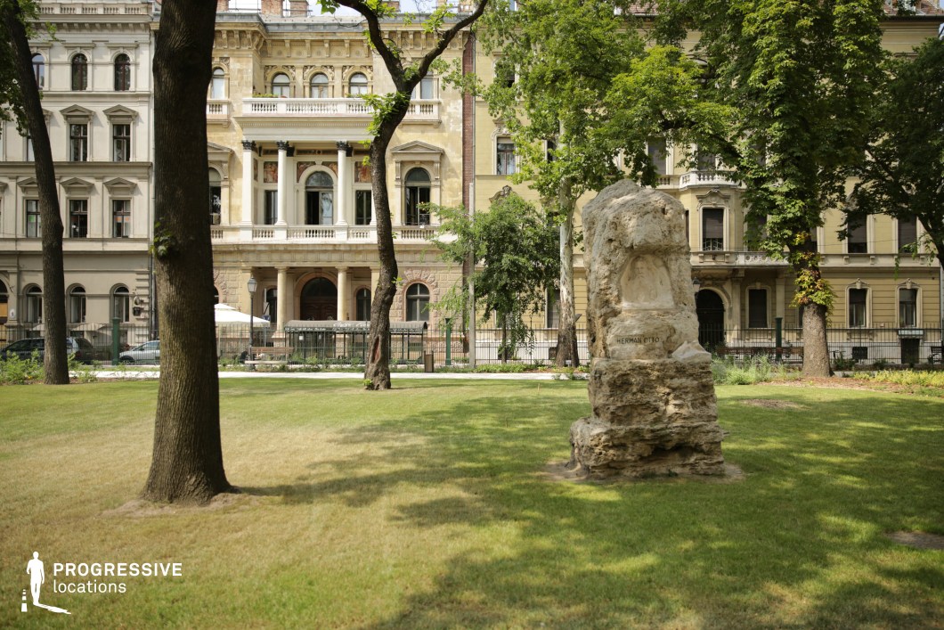 This serene park scene features a prominent stone statue, dedicated to Herman Otto, placed on the lush green lawn. The statue, slightly weathered, stands in sharp contrast to the modern well-maintained grass, creating a sense of history within the space. In the background, classical buildings with intricate facades rise above the park, providing an architectural backdrop that complements the statue's historical significance. Tall trees line the edges of the park, further framing the scene, creating a peaceful environment that balances both nature and heritage. The combination of the monument, the well-preserved parkland, and the elegant buildings creates a sophisticated setting, making this location ideal for cinematic scenes with themes of history, reflection, and urban beauty. The contrast between the stone statue and the surrounding modern greenery allows for visually rich compositions, perfect for wide shots or detailed close-ups of both the statue and the surrounding architecture.