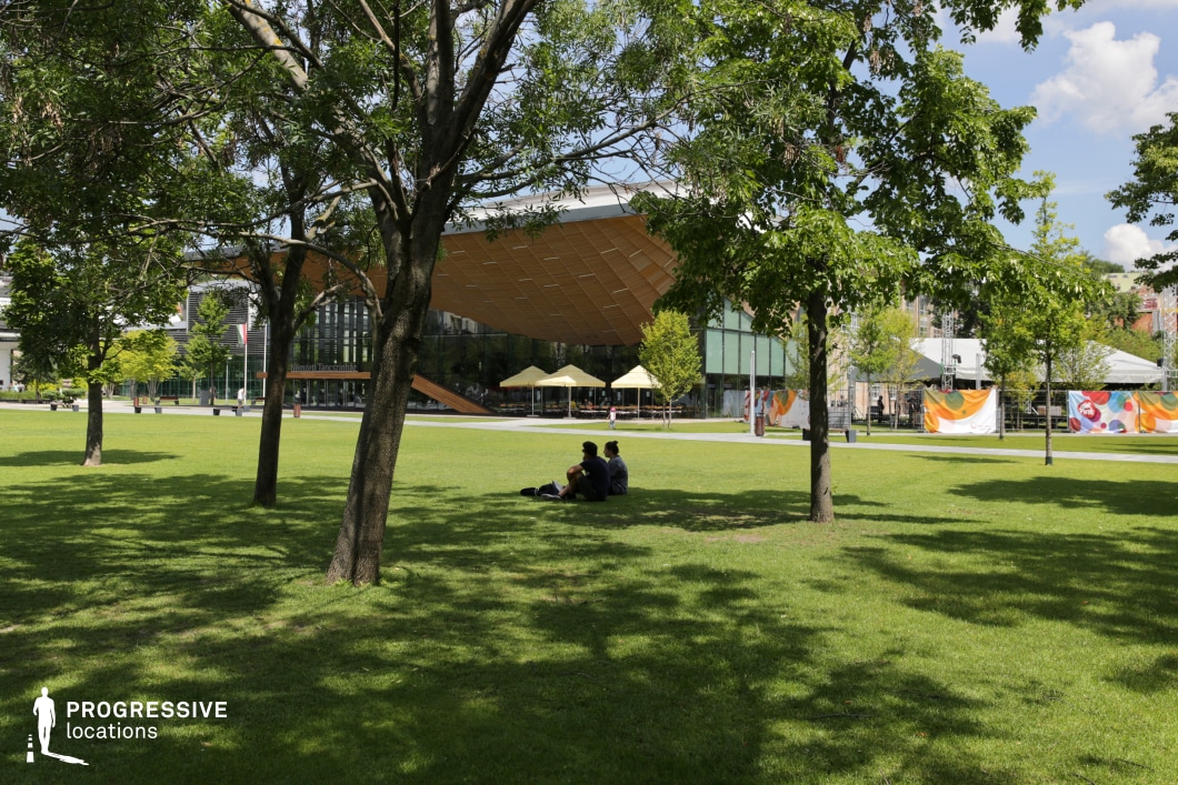 This modern park scene features a large, open green space with well-manicured grass, bordered by rows of young trees providing both shade and natural framing. The sleek, contemporary architectural pavilion in the background adds a dynamic focal point to the scene, with its bold, angular roof and glass facade creating a striking contrast against the greenery. The pavilion is surrounded by minimalistic, modern elements, including shaded seating areas and temporary event structures. This location offers a versatile setting, ideal for both wide shots capturing the harmony between nature and modern architecture, and more intimate close-ups of the pavilion's design features. The park's open layout and the pavilion’s clean lines make it perfect for a variety of cinematic themes, from contemporary urban life to design-focused narratives. The blend of natural beauty and modern structures provides an ideal environment for dynamic, visually appealing scenes.