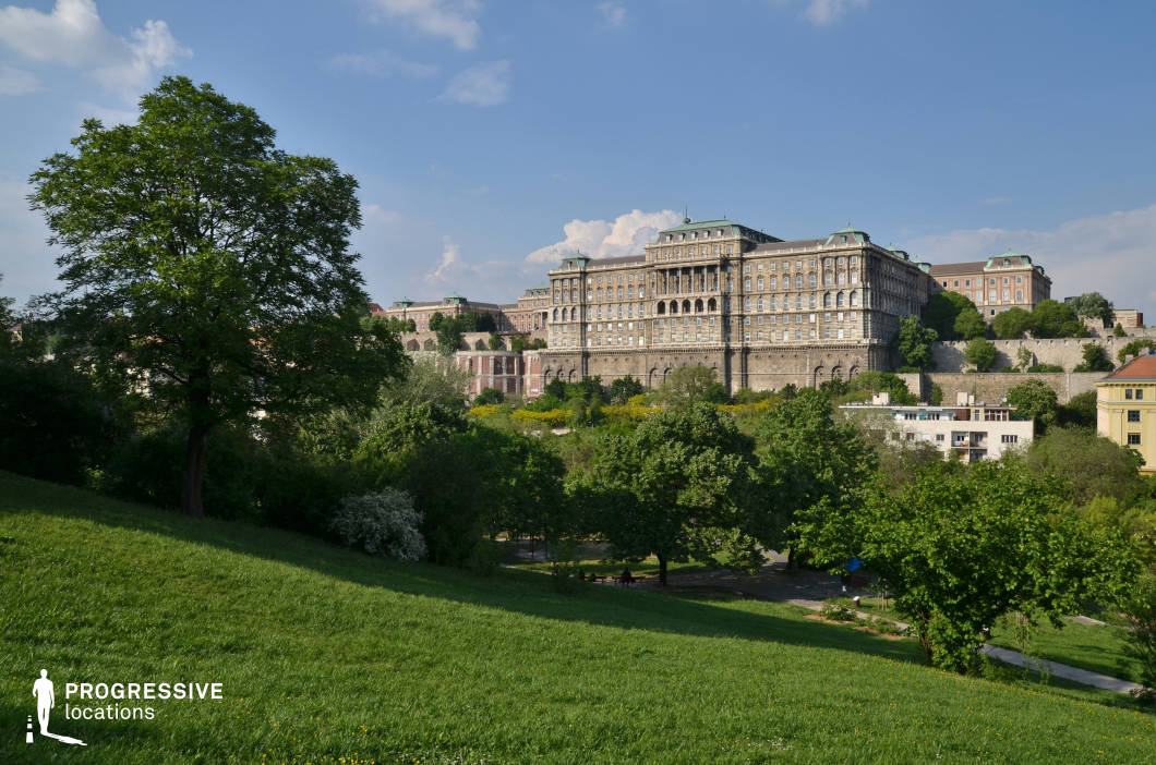 This image showcases a gently sloping park pathway leading towards a stunning panoramic view of a grand historic castle. The pathway is surrounded by lush greenery, with mature trees casting dappled shadows on the pavement. Well-maintained grassy areas and neatly trimmed bushes frame the scene, enhancing the park’s inviting and natural atmosphere. In the background, an imposing neoclassical-style castle with intricate architectural details and a domed rooftop dominates the skyline. The castle’s stately design and elevated position contribute to the sense of grandeur and historical significance. The combination of parkland and historic architecture creates a visually rich contrast between nature and urban heritage. This location is ideal for cityscape photography, historical-themed productions, or scenic lifestyle imagery, capturing both natural beauty and architectural magnificence in one frame.