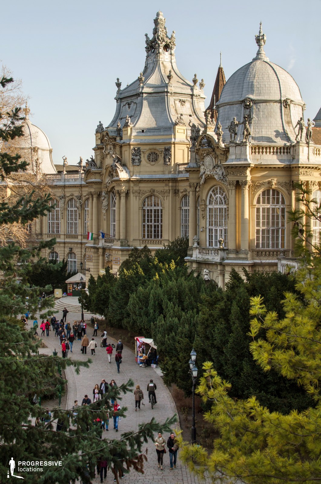 This striking image showcases a grand baroque-style building, featuring ornate domes and intricate stonework on its facade. The architecture is adorned with classical elements, such as detailed statues and expansive windows, giving it a majestic and historical atmosphere. In the foreground, a smooth, well-paved pathway leads towards the entrance, framed by lush greenery on either side. The combination of the architectural grandeur and the natural surroundings creates a harmonious contrast, enhancing the visual appeal of the space. This location is perfect for film scenes that require a sophisticated historical backdrop, offering both the elegance of baroque design and the tranquility of a well-maintained park setting. The path and architectural details provide ample opportunities for dynamic cinematic shots, whether wide-angle views of the building or focused close-ups of the intricate elements.