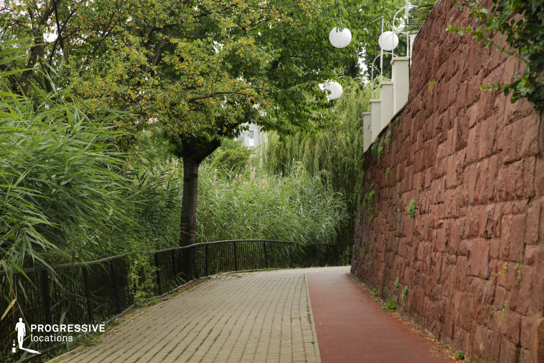 This tranquil scene captures a smooth pedestrian pathway, bordered by a tall stone wall on one side and dense greenery on the other. The pathway is well-maintained, with a clean, paved surface and a gentle curve, leading the eye through the lush environment. On the left, vibrant green reeds and trees provide a natural buffer, creating a sense of seclusion. The stone wall, with its textured red hues, adds a rustic, architectural element, offering contrast to the natural surroundings. The presence of white lanterns mounted on the wall introduces a modern touch, suggesting a design that blends tradition with contemporary aesthetics. This location is ideal for film scenes that require a peaceful, atmospheric setting, perfect for moments of solitude or contemplation. The balance of organic elements and structured design creates visual interest, making it suitable for wide-angle shots or focused close-ups that highlight the interplay of nature and architecture.
