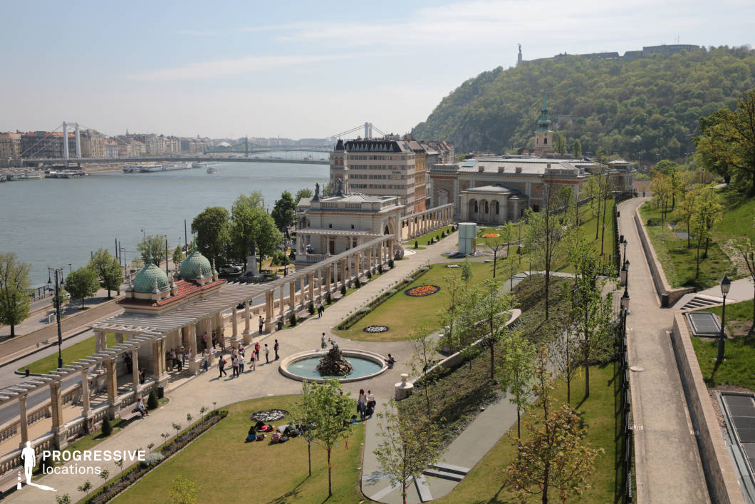 This panoramic view captures a beautifully landscaped riverside park, bordered by historical buildings and framed by the calm waters of the river. The scene showcases a wide pathway that meanders along the riverbank, with a row of stone columns and a majestic pergola adding classical architectural charm. The central fountain, adorned with intricate sculptures, adds a focal point to the well-kept greenery, while flowerbeds enhance the aesthetic appeal. In the distance, the city’s buildings rise against the backdrop of the rolling hills, offering a seamless blend of urban and natural landscapes. The elevated viewpoint provides a sense of grandeur and depth, ideal for cinematic shots requiring sweeping views. The park’s combination of refined architecture, open green spaces, and proximity to the water creates a versatile setting for various film genres, from period dramas to romantic or scenic scenes. This location offers a dynamic environment, perfect for both wide shots and detailed close-ups, with its mix of historical and natural elements allowing for diverse visual storytelling.