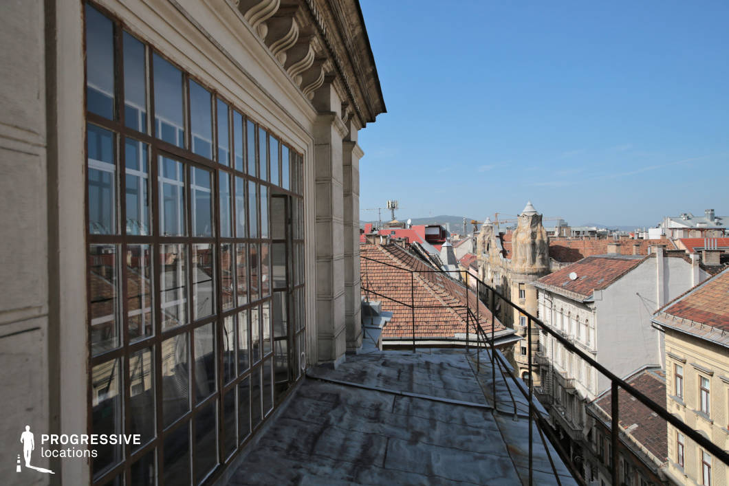 This image presents a view from a terrace that extends beyond a classical architectural building, offering a distinctive perspective of the city rooftops. The metal railing adds a contemporary touch, contrasting with the ornate detailing of the building's facade, visible just next to the large multi-pane window. The building’s window reflects parts of the surrounding rooftops, showing a mix of red-tiled and flat roofs, as well as the unique, rounded turret of a neighboring building, which contributes a sense of historical charm. The scene conveys an open, airy feeling, emphasized by the clear blue sky. The space itself appears spacious, offering ample room for an outdoor setting. The juxtaposition of old and new architectural elements makes this a versatile location for both contemporary and period-inspired shoots, adding an urban, timeless atmosphere with views that stretch across the city. This terrace could serve well for urban lifestyle photography, architectural shoots, or settings that call for a high vantage point in an urban environment.