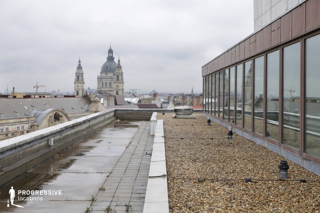 This image showcases a rooftop scene where a modern glass structure is reflected in the windows of a building. The foreground presents a flat, gravel-covered roof with minimal vegetation, slightly wet, suggesting recent rainfall. The backdrop features a striking view of a large basilica with two prominent towers rising above the surrounding cityscape. The dome of the basilica is clearly visible, adding a historical and architectural focal point to the scene. The muted, cloudy sky creates a somber and atmospheric mood, enhancing the visual contrast between the old architecture of the basilica and the modern building with its clean glass panels. The combination of these elements offers a unique juxtaposition of the old and new, ideal for visual storytelling involving cityscapes or urban contrasts. The horizontal lines of the rooftop and the reflections in the glass contribute to the scene’s clean, minimalistic composition, which could be used for contemporary urban-themed photo shoots or architectural explorations.