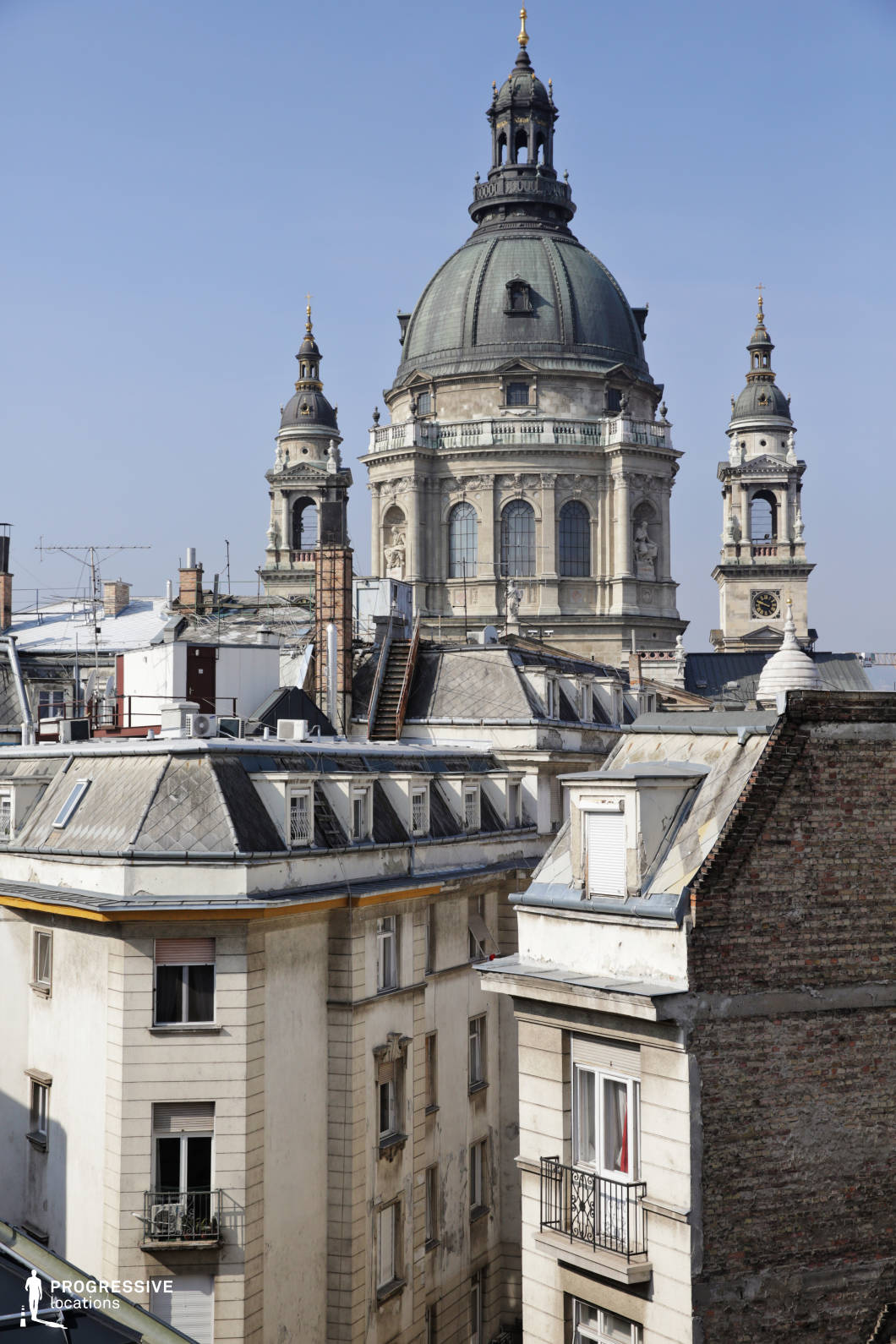 This image features a close-up of a grand basilica dome set against the backdrop of surrounding rooftops. The basilica's large, domed roof is framed by the detailed spires of its towers, showcasing intricate stonework and large arched windows. The rooftops in the foreground are a mix of aged buildings with steeply sloped roofs, featuring dormer windows and various rooftop accessories like antennas and air conditioning units. The cityscape is filled with a sense of historical depth, as the classical architecture of the basilica contrasts with the more utilitarian, residential rooftops. The clean, bright blue sky enhances the prominence of the dome and the sharp details of the surrounding rooftops, creating a visually striking image that combines both grandeur and the daily life of the city. The composition allows for an intimate view of the urban environment, offering potential for architectural photography or urban-themed shoots that highlight both the old-world charm and modernity of the space.