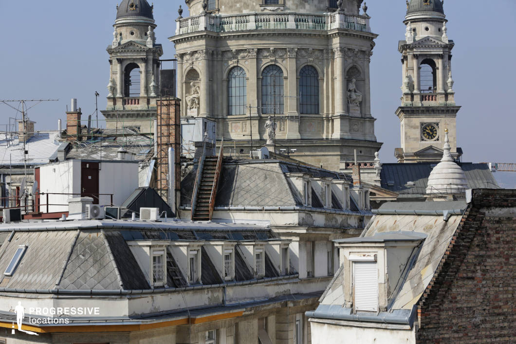 This image offers a detailed view of rooftops with the prominent basilica towers rising above. The foreground captures a variety of sloped, tile-covered roofs with various rooftop elements such as dormer windows, antennas, and air conditioning units, creating a densely packed, functional urban landscape. The basilica’s towering structure stands out in the background, with its intricate stonework and large, arched windows. The clock tower, part of the basilica, is framed in the composition, providing a strong vertical element against the horizontal expanse of rooftops. The detailed carvings on the basilica’s façade, especially the statues near the windows, enhance the historical character of the scene. The bright blue sky creates a vivid contrast, making the scene feel open and expansive while emphasizing the grandeur of the basilica. This location, rich with architectural contrast between the modern rooftops and the historical basilica, is perfect for shoots exploring the tension between old and new, ideal for architectural photography or urban-centric projects.