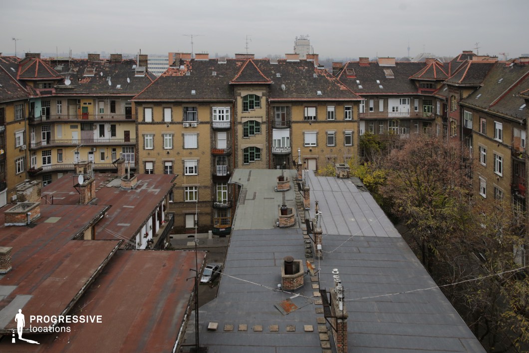 This image offers a view of rooftops and apartment buildings from an elevated angle, creating a sense of intimacy with the surrounding urban environment. The rooftops, covered with a combination of corrugated metal and tiles, are visible from above, with various chimneys and vents adding texture to the scene. The buildings themselves are made of brick, their facades characterized by rectangular windows and small balconies, offering a sense of residential life. The courtyard between the buildings is lined with trees, some of which show signs of having lost their leaves, contributing to the scene's somewhat muted, autumnal atmosphere. The light gray sky sets a somber tone, complementing the earthy tones of the buildings and rooftops. The scene offers a glimpse into a quiet urban setting, ideal for capturing the essence of city living or exploring themes of urban architecture. The visual layout of the rooftops provides an interesting backdrop for photography, highlighting the architectural uniformity and occasional contrast found in densely built environments.