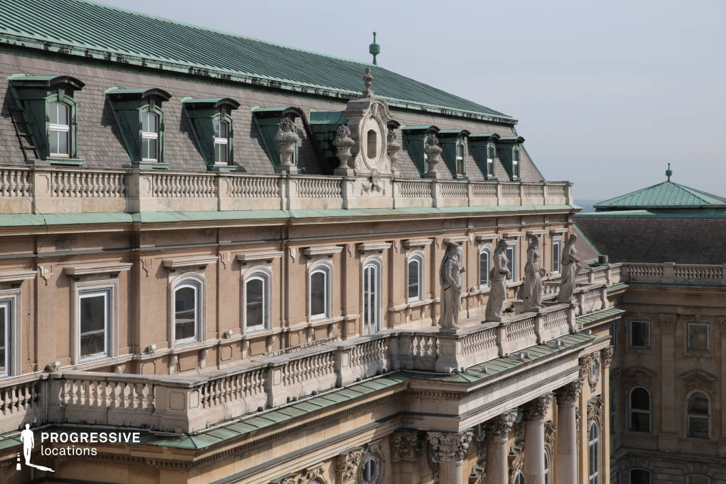This image highlights an intricate detail of the Buda Castle’s rooftop, focusing on the elegant architecture and statuary that adorn the building’s upper level. The green patina on the roof’s copper elements contrasts with the warm beige stone of the building’s façade. The statuary along the terrace railing includes finely sculpted figures, adding a classical touch to the structure, while the detailed balustrade reinforces the grandeur of the space. The windows, framed with ornamental moldings, add symmetry and depth to the composition. The sky is clear, allowing the rooftop’s rich textures and the ornate elements to stand out. The overall feel is one of historic elegance, showcasing the architectural refinement of the Buda Castle. This setting is perfect for projects that require a high-class, historical location with stunning architectural details, ideal for both cinematic and photographic purposes focused on grandeur, heritage, and cultural beauty.