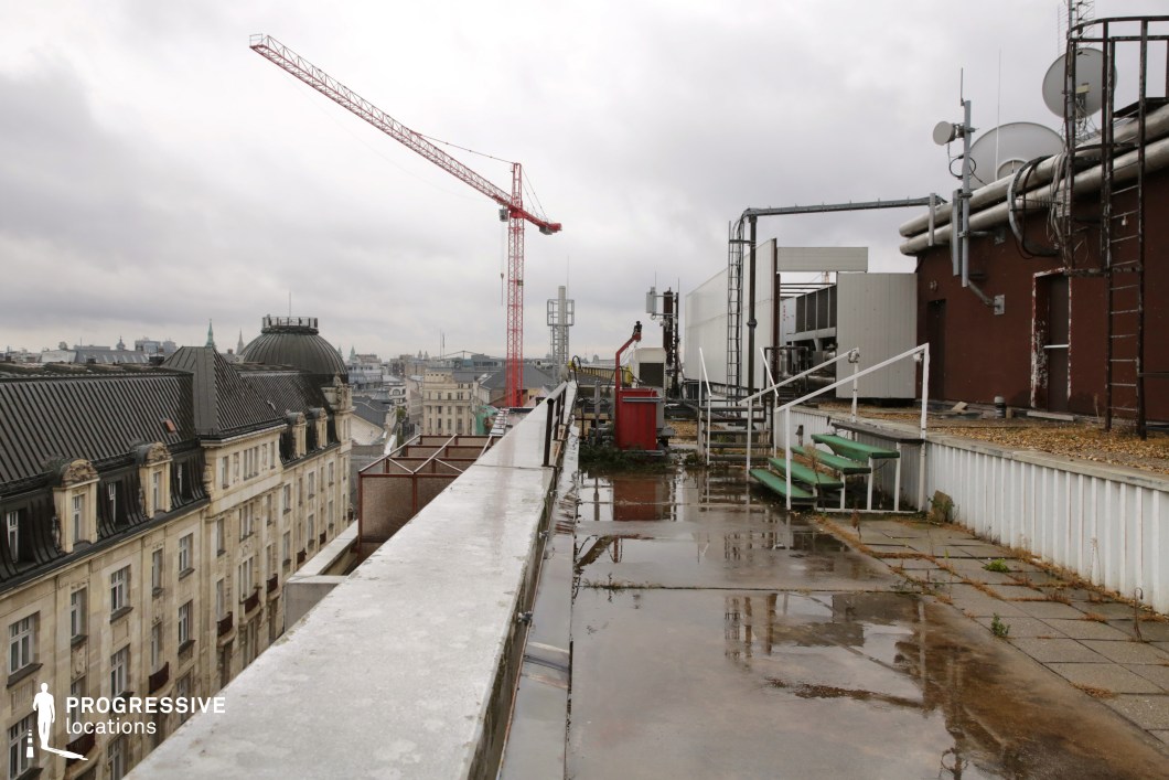 This image presents a rooftop with a clean, paved surface that overlooks a sprawling cityscape. The rooftop is surrounded by various industrial elements such as air conditioning units and metal staircases, adding a modern, functional aspect to the setting. In the background, the city’s skyline unfolds, featuring a mix of older, classical buildings with a prominent dome-shaped structure and more contemporary high-rise buildings in the distance. The overall atmosphere is one of urban vibrancy, with a contrast between the historical architecture and the industrial rooftop. The clear sky provides a bright, open backdrop, emphasizing the vastness of the city below. This location is ideal for capturing scenes focused on the blend of old and new architecture, offering a versatile backdrop for urban photography or city-themed shoots.