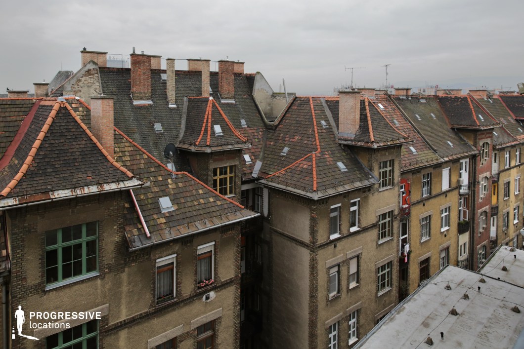 This image presents a view of several rooftops within a residential urban environment, showcasing the intricate design of slanted roofs with red tiled accents. The buildings, with their warm-toned brick façades, exhibit a mixture of colors, from yellow to brown, adding visual depth to the scene. The rooftops are adorned with chimneys, skylights, and antennas, contributing to the city’s characteristic architectural rhythm. A subtle contrast in textures is visible, as some buildings display smoother surfaces while others reveal more weathered brick and exposed architectural details. The muted sky complements the overall somber, urban atmosphere, highlighting the density and compactness of this neighborhood. This location is ideal for capturing the essence of city life, with a focus on residential architecture and the architectural complexity of older buildings. It is well-suited for urban photography or projects that emphasize the texture and structure of city environments.