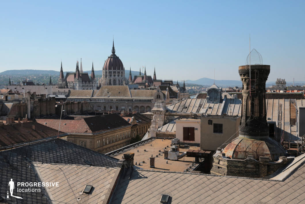 This image captures a dynamic rooftop view that highlights the blend of old and new architecture in the city. In the foreground, several rooftops are visible, characterized by their distinct materials, such as sloped tiles and flat roofs. Among these rooftops, various industrial elements like chimneys and ventilation systems add a utilitarian touch to the scene. In the background, the grand Hungarian Parliament building rises majestically, its intricate spires and detailed façade standing out against the surrounding cityscape. The clear sky creates a sharp contrast with the architectural details, enhancing the historical grandeur of the Parliament. The hills in the distance provide depth and a scenic backdrop to this vibrant urban setting. This location is ideal for capturing the contrast between the classical elegance of the Parliament building and the modern, functional rooftops of the city, offering a unique perspective for urban photography and architectural-focused projects.