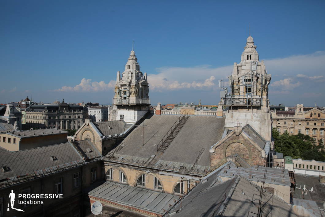 This image offers a rooftop view that emphasizes the intricate architecture of two prominent towers, standing out against a backdrop of a sprawling cityscape. The towers, adorned with detailed stonework and decorative elements, dominate the skyline with their distinct profiles. The scaffolding surrounding the towers hints at ongoing restoration work, contrasting with the historical architecture's grandeur. Below, the rooftops display a blend of old, functional buildings with sloped metal roofs, creating a textured, urban landscape. The open, clear sky above enhances the visual contrast between the restored, classical features of the towers and the more utilitarian elements of the surrounding buildings. This scene captures a vibrant yet historical urban atmosphere, making it ideal for architectural photography, cityscape shots, or projects focused on the preservation and development of heritage buildings.