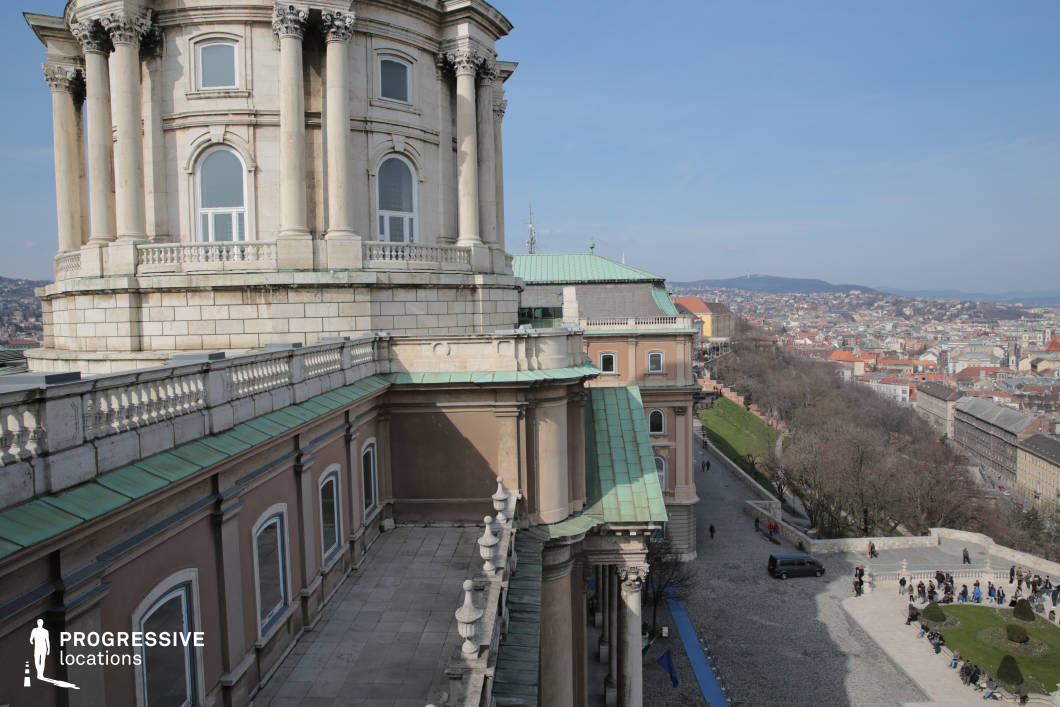 This image offers a stunning view from the terrace of a historic building, possibly a national gallery, with the dome and columns in the foreground. The smooth, light-colored stonework of the building is contrasted by the copper roof, showing signs of patina. The terrace’s stone railing provides an elegant border, with decorative finials accentuating the classical architecture. In the distance, the city unfolds, with its mix of rooftops, green hills, and the expansive urban sprawl. The scene is bathed in bright, natural light, with the clear blue sky enhancing the open, airy feeling of the space. Below, the wide cobbled walkway leads to a lush, green park, where people can be seen enjoying the outdoors. This location, with its sweeping views and architectural elegance, is perfect for capturing the grandeur of both the building and its surroundings, offering a rich backdrop for urban or historical photography. It embodies a blend of cultural heritage and the beauty of nature in the city.