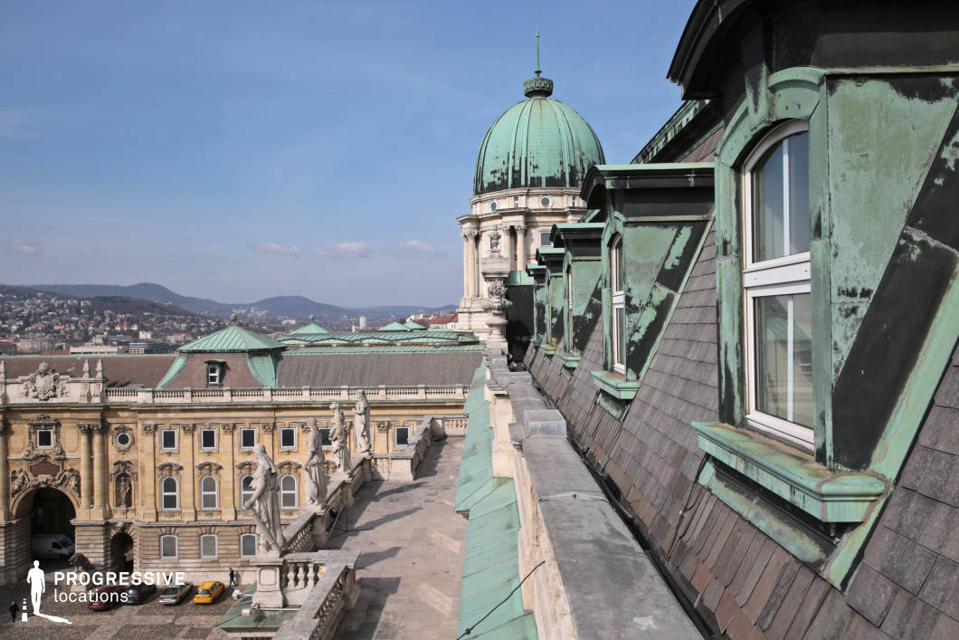 This image offers a close-up view of a mansard roof adorned with weathered copper elements. The roof is sharply angled, with several dormer windows and metal detailing that reflect the passage of time. In the distance, the prominent dome of the building stands out, featuring its distinctive green patina, which contrasts with the warmer tones of the surrounding architecture. Below the roofline, the architectural grandeur of the building is enhanced by sculptural elements, including statues lining the terrace. The city’s skyline, with its undulating hills, provides a picturesque backdrop to this historical location. The bright, clear sky above adds vibrancy, further highlighting the detailed craftsmanship of the dome and roof. This scene is ideal for capturing the blend of architectural elegance and urban landscape, making it a suitable setting for both architectural photography and historical documentation. The combination of textures and the commanding view of the city below creates a compelling composition full of depth and visual interest.