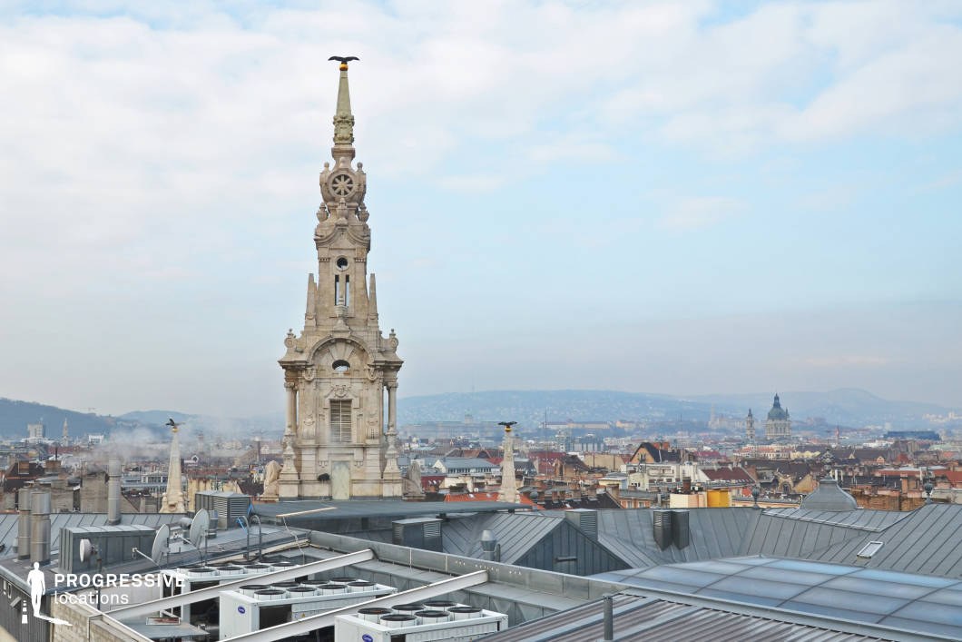 This image captures a unique rooftop perspective with a prominent stone tower in the foreground. The tower, adorned with intricate architectural details, rises high above the surrounding rooftops, with a bird perched atop its spire. Below, the rooftops display a mix of modern and traditional elements, with metallic and flat roof structures contrasting with the older stone buildings. The panoramic cityscape stretches into the distance, with distant hills and iconic landmarks visible, including domes and spires that punctuate the skyline. The soft, hazy sky adds a tranquil ambiance to the scene, emphasizing the grand scale of the tower against the sprawling urban environment. This location is ideal for capturing the dynamic relationship between historical and modern elements in a city, making it suitable for both architectural and urban photography projects that explore the contrasts within cityscapes.