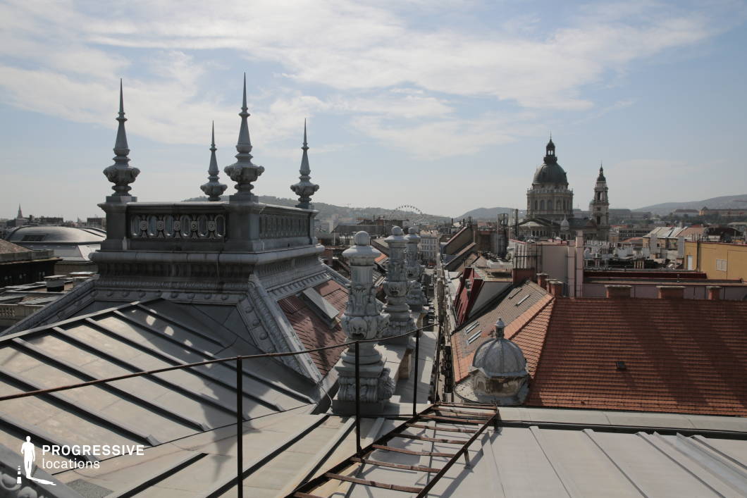 
This image captures a view from a rooftop featuring decorative metal spires that stand out against the city backdrop. The detailed craftsmanship of the roof’s design is evident in the ornate spires, creating a sense of historical grandeur. In the background, the iconic basilica dome rises prominently, flanked by a tall tower, adding depth and focus to the scene. The combination of sharp architectural features and the smooth, angled rooftops below creates an interesting contrast between the intricate and the functional. The expansive cityscape stretches out in the distance, with rolling hills and distant structures visible under the bright, clear sky. This scene perfectly embodies the blend of old-world architectural beauty and urban life, offering a unique perspective of a historic city. The location is ideal for capturing the dynamic relationship between architectural elements, suitable for urban or architectural photography.