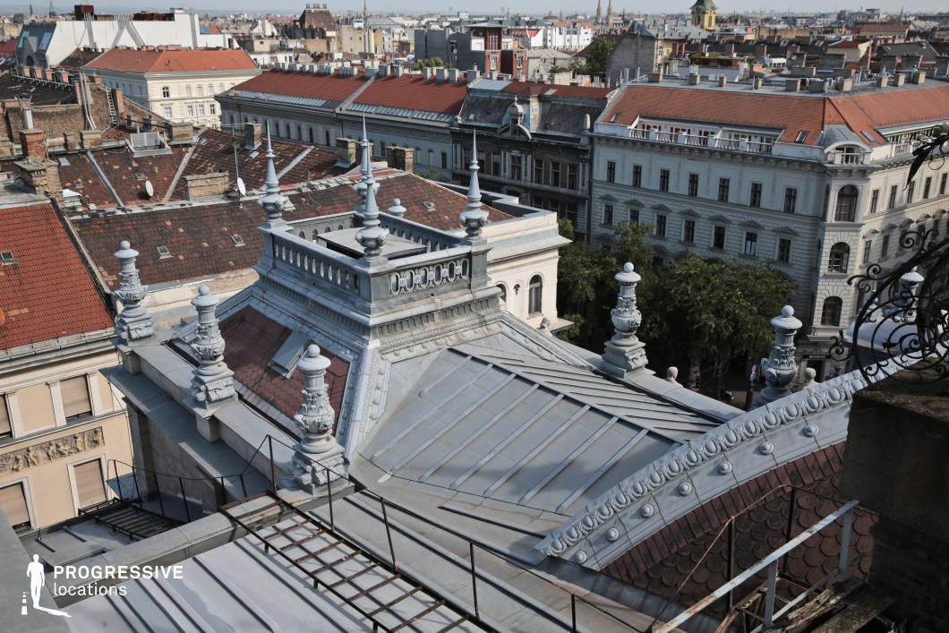 This image offers a detailed view of a rooftop adorned with ornate metalwork and decorative spires. The intricate design of the roof, with its mix of copper elements and tiled surfaces, reflects the building’s historical elegance. Below the rooftop, the architecture of the surrounding buildings on Andrassy Avenue is visible, with their classical facades adding depth and character to the scene. The sharp spires and stately chimneys of the rooftops create a distinctive silhouette against the city skyline, while the Opera House can be seen in the distance, contributing a sense of cultural grandeur to the composition. The green trees lining the street below provide a lush contrast to the urban environment. The bright sunlight enhances the fine details of the architecture, showcasing the meticulous craftsmanship of the building’s design. This location, with its fusion of cultural heritage and urban landscape, is perfect for projects centered around architectural elegance or capturing the charm of Budapest’s historical cityscape.