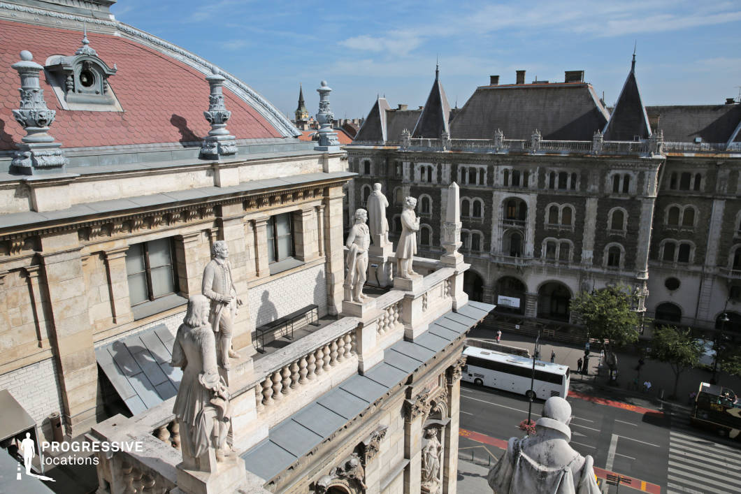 This image captures the rooftop terrace of the Opera House, where a series of statues elegantly line the building's façade. The figures, carved in marble, stand tall against the detailed architecture, adding a classical touch to the structure. The statues are positioned along a decorative balustrade, providing a sense of grandeur and history. In the background, the rooftops of adjacent buildings are visible, showcasing a mix of traditional and more modern designs. The vast cityscape stretches beyond, with urban streets and traffic visible below, adding a dynamic contrast to the serene and monumental nature of the rooftop terrace. This location is perfect for capturing the blend of historical architectural beauty with the lively energy of the surrounding city. The scene is ideal for projects that explore both the grandeur of cultural landmarks and the contrast of urban life.