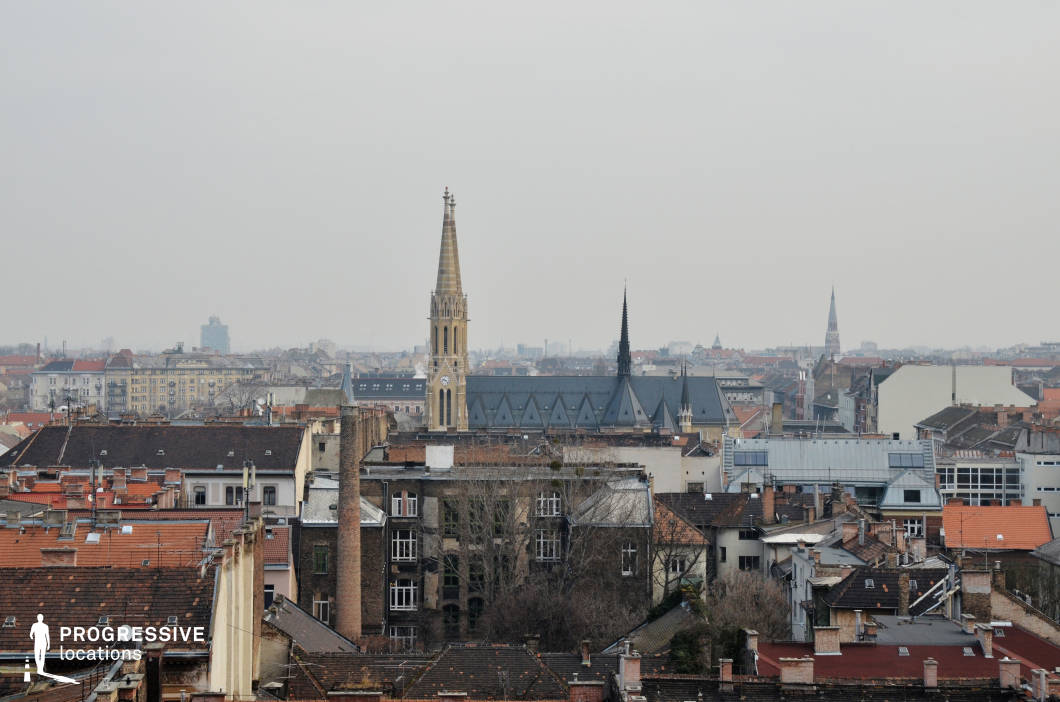 This image captures a panoramic view of a city’s rooftops, with a striking focus on the towering church spires that dominate the skyline. The rooftops are densely packed, featuring traditional tiled roofs and a variety of architectural styles that reflect the historical charm of the area. In the foreground, a tall, elegant church tower stands out with a clock, while another spire rises alongside it, providing vertical contrast to the horizontal expanse of the surrounding rooftops. The scene is framed by a mix of older, classic buildings and more modern structures, creating a harmonious blend of architectural eras. This location offers a picturesque setting for capturing the essence of urban life, with a particular emphasis on religious or historical architecture, making it ideal for projects that explore the evolution of city landscapes.