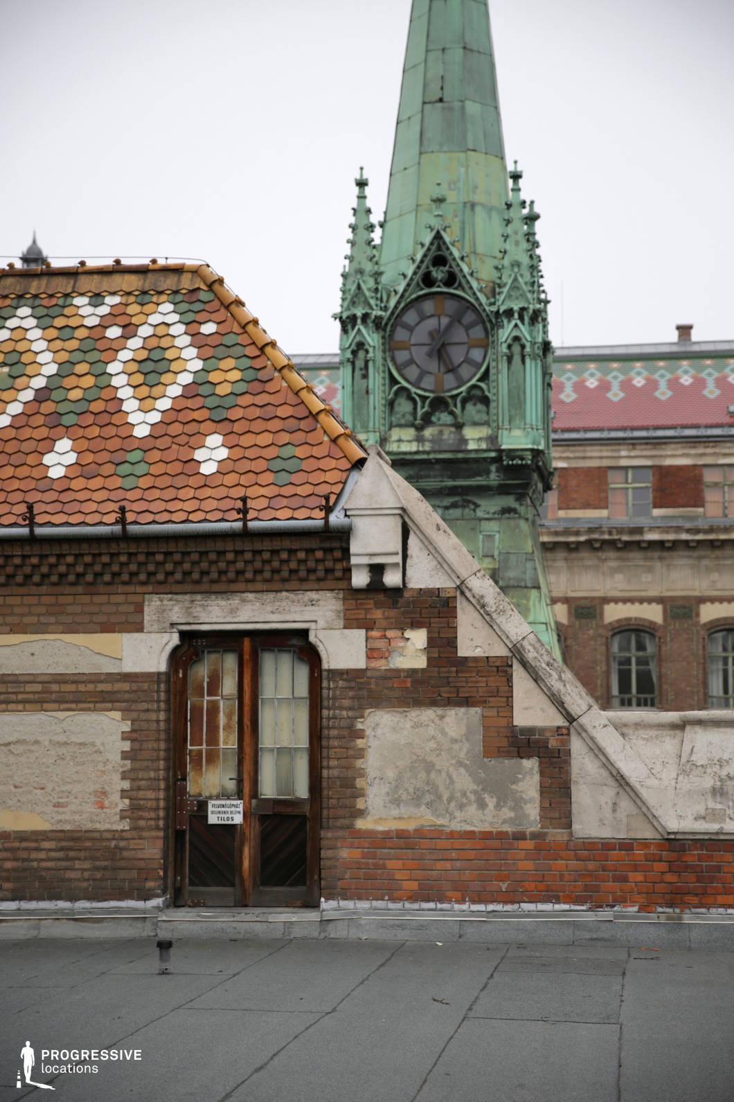 This image captures the distinctive details of a university library rooftop, featuring a decorative tiled roof with a blend of warm colors in hexagonal shapes. The roof, complemented by the textured brick walls and an old wooden door with a vintage sign, forms a charming yet weathered part of the structure. In the background, a tall clock tower rises, its green copper spire and ornate clock face adding an impressive focal point to the scene. The architectural elements reflect a combination of classical and historic design, with worn finishes that speak to the age and history of the building. This location offers a compelling view that highlights the balance between historical architectural beauty and the evolving urban environment. The clock tower and tiled roof make it a perfect setting for projects focused on heritage, academic spaces, or architectural photography.