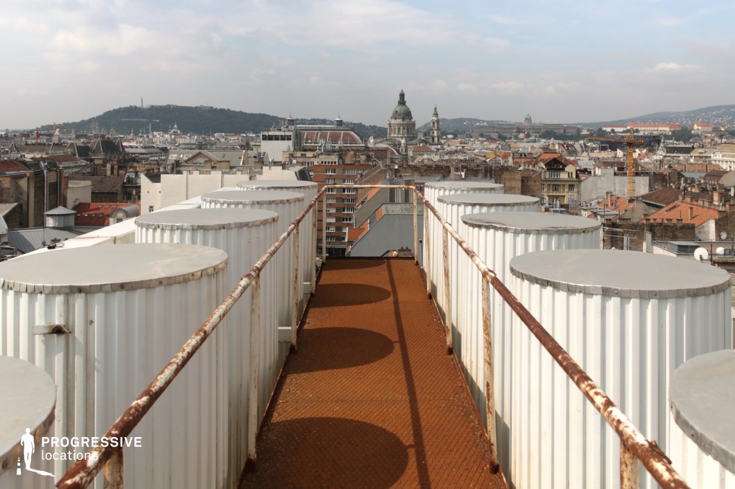 This image presents a unique rooftop view featuring an industrial corridor lined with large cylindrical tanks. The rusted railing and textured flooring provide a gritty contrast to the otherwise clean and organized cityscape in the background. The city is dominated by rooftops and the iconic dome of a basilica, with distant hills framing the horizon. This juxtaposition of industrial elements and historic architecture highlights the blend of functional, modern infrastructure with the charm of older urban landscapes. This scene is ideal for capturing the interaction of industrial elements within the context of an evolving city environment. Perfect for projects that explore the intersection of architecture, urban renewal, and the utilitarian side of city life.