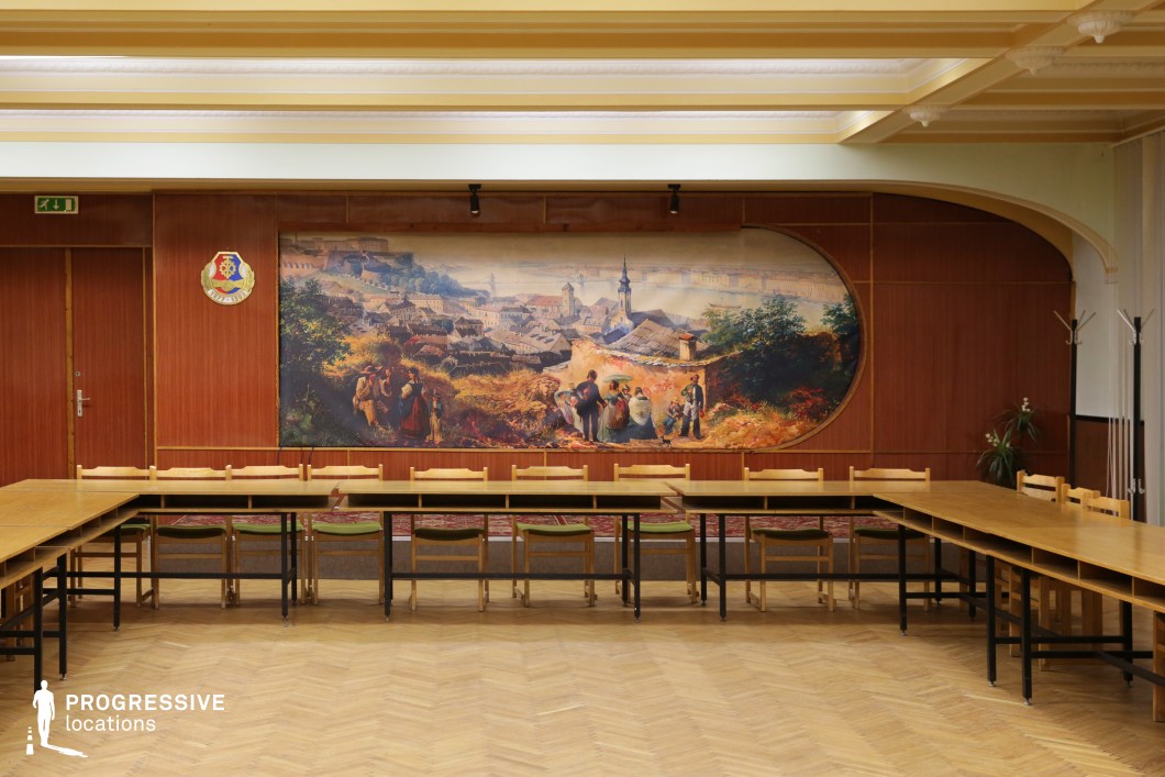 This image showcases an interior space designed for formal or professional settings, featuring a large mural in the background. The room is furnished with a series of long wooden tables arranged to form a spacious, rectangular seating arrangement. The floor is made of light wooden parquet, enhancing the room's clean, organized aesthetic. In the background, a large historical mural adds a classical touch, depicting a rural scene with figures in traditional attire, subtly blending the natural environment with the architectural elements. The wood paneling and soft lighting further highlight the warm and formal atmosphere of the room. The coat of arms mounted on the wall adds a touch of distinction, suggesting that the room is used for official or civic purposes. The layout of the tables and chairs creates a functional space that could accommodate meetings or gatherings, emphasizing its suitability for formal discussions, debates, or organizational activities. This versatile setting can serve a variety of functions, making it ideal for professional events, historical-themed presentations, or community gatherings.