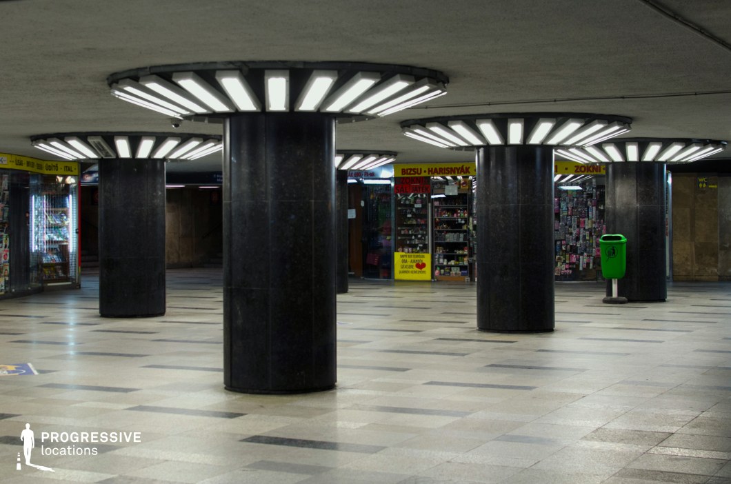 This image presents a modern underground passage with a functional, utilitarian design. The space is characterized by large, dark columns topped with circular fluorescent lighting, which creates a stark, industrial atmosphere. The floor is made of symmetrical, checkered tiles that reinforce the clean, organized aesthetic of the area. In the background, vending kiosks line the passageway, suggesting the area's role as a transit hub or commercial space. The vibrant green trash bin adds a contrasting touch of color amidst the neutral tones of the space. The lighting and architectural elements contribute to a well-maintained and practical environment, ideal for high-traffic urban scenes or functional settings in film and photography. The layout is open and spacious, allowing for dynamic movement or interaction, making the location versatile for various creative projects.