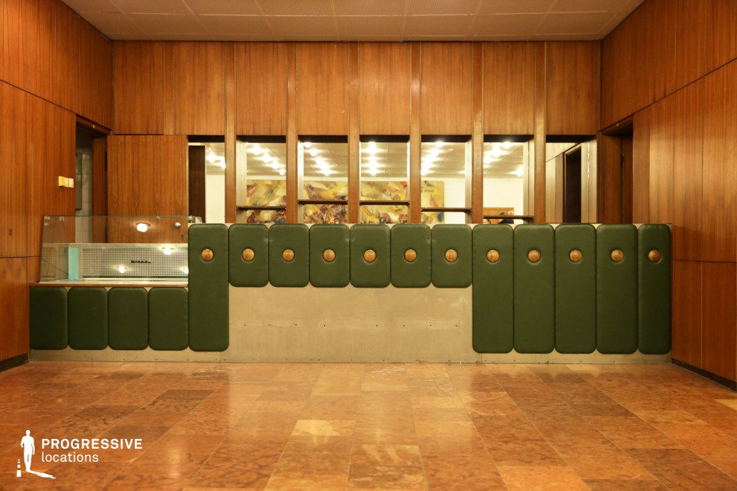 This image features a stylish bar counter with a distinctive mid-century modern design. The counter is made of smooth green leatherette panels, each with circular brass buttons, creating a rhythmic, geometric pattern across the surface. The base of the counter is finished with a sleek, light-colored stone or concrete, providing a contrasting texture. Behind the counter, wooden paneling extends along the walls, contributing to the warm, inviting atmosphere. The room beyond the counter is softly illuminated, with indirect lighting creating a relaxed yet refined ambiance. The polished marble floor, with its rich reddish-brown tones, complements the luxurious feel of the space. This setup could serve as an elegant, retro-inspired location for professional or social events, lending a timeless, upscale atmosphere to any scene. The functional and visually striking design elements make it adaptable for a variety of creative uses, from historical reenactments to modern lounge scenes.