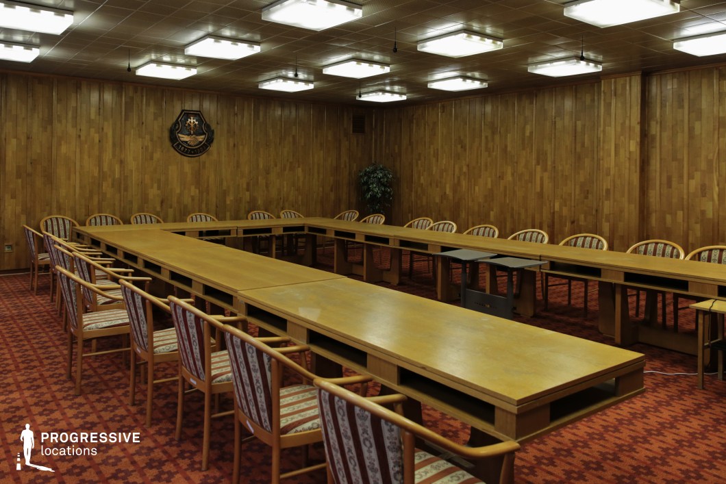 This image presents a formal boardroom setting, characterized by a long rectangular table surrounded by wooden chairs, all set up for a professional gathering. The walls are clad in light wood paneling, contributing to the room's warm, traditional atmosphere. The red carpet flooring, with its intricate pattern, adds to the vintage charm of the space. Above, the ceiling is evenly illuminated by square light fixtures, ensuring a well-lit environment suitable for meetings or conferences. At the far end of the room, a coat of arms is displayed, suggesting the official nature of the setting. The room’s symmetrical layout, along with the neatly arranged furniture, emphasizes the space’s formality, making it ideal for corporate discussions, formal meetings, or any professional setting where authority and decorum are paramount. This location can serve as a backdrop for scenes requiring a traditional, official environment or for any narrative needing a serious, controlled setting.