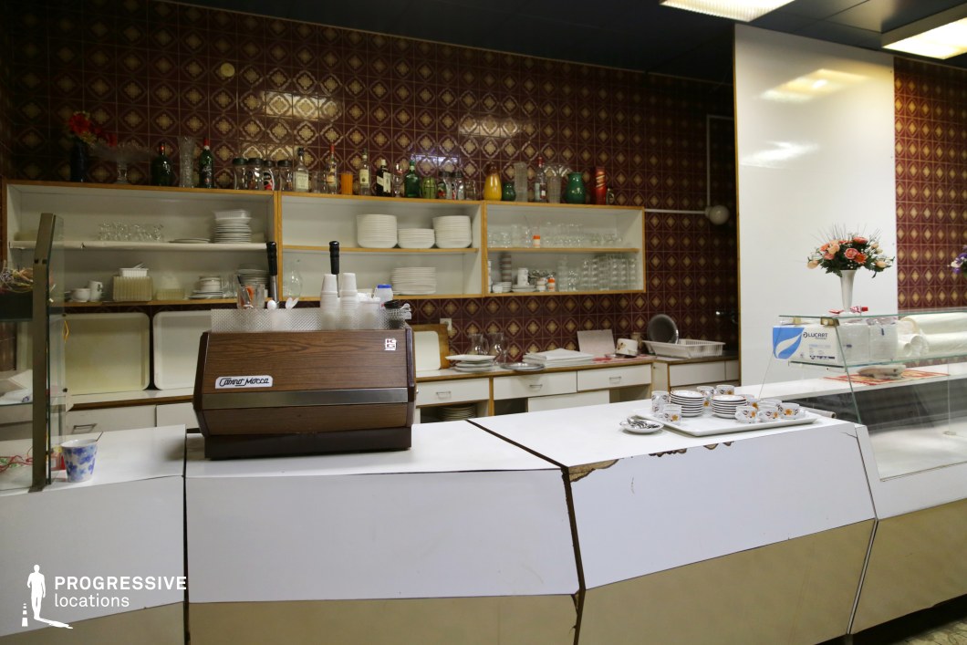 This image presents a vintage-style buffet counter, featuring a white and brass exterior with noticeable wear along the edges, suggesting its age. The counter is partially enclosed with a glass display section, showcasing plates with floral patterns, a nod to retro dining aesthetics. Behind the counter, a wall is covered with patterned burgundy tiles, which adds a nostalgic, warm tone to the space. Shelves above the counter hold a collection of glasses, bottles, and dishes, including traditional glassware and vintage bottles. The setting is decorated with a small flower arrangement on the counter, adding a subtle, homely touch. The overall ambiance of the room conveys a charming, old-fashioned feel, making it ideal for scenes set in classic diners, retro-themed establishments, or nostalgic moments in film, where the setting evokes memories of past decades or simpler times. This location is also perfect for portraying everyday life in a more utilitarian, yet nostalgic, environment.