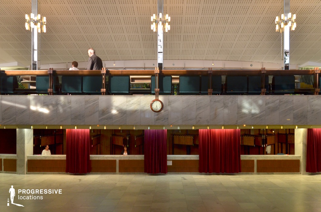 This image captures the interior of a grand theatre lobby, featuring an open space with clean, modern lines. The upper level is lined with deep green seating, overlooking the main floor, which is marked by a sleek marble counter beneath a row of red velvet curtains. The counter is separated by these curtains, which create private sections for cloakroom or ticket services, enhancing the sense of exclusivity. A large clock, mounted on the wall above, adds a classic touch, while the softly lit chandelier casts a warm glow over the scene. The spacious design and elegant finishes suggest the theatre’s high-end, cultural atmosphere, making this location ideal for formal gatherings, performances, or high-profile events. This environment is perfect for capturing moments of transition or anticipation, offering an excellent backdrop for cinematic scenes in settings that require grandeur or sophistication.
