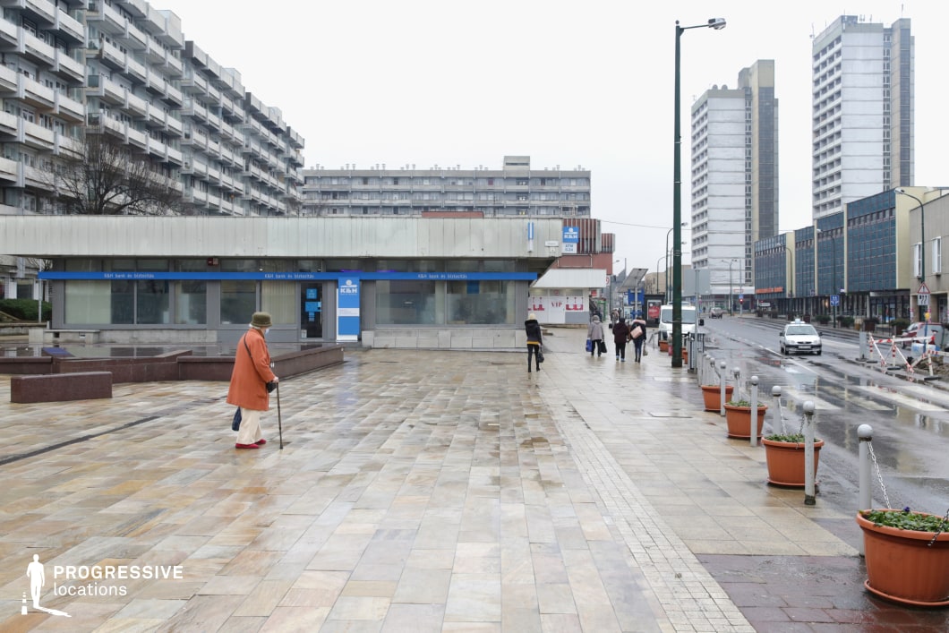 This image captures a modern urban plaza with Brutalist architecture in the background. The scene features wide, paved walkways made of large square tiles, leading toward a bank building with a sleek, minimalist design. A single elderly figure walks across the plaza, dressed in a long orange coat and red shoes, adding a human element to the otherwise open and quiet space. Tall concrete apartment buildings with repetitive, angular designs dominate the skyline, characteristic of Brutalist and post-war urban planning. The surrounding area includes street lamps, planters with small trees, and some pedestrians, contributing to the functional and pragmatic feel of the location. This scene is ideal for depicting city life, urban landscapes, or residential areas with a focus on modernist, utilitarian design. The layout and muted colors provide a versatile backdrop for various cinematic or commercial uses, highlighting everyday moments in a stark, urban environment.