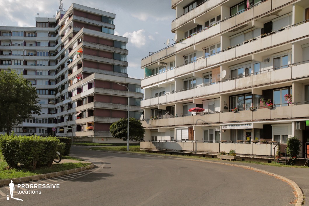 This image features a Brutalist residential complex, showcasing several multi-story apartment buildings with angular, exposed concrete facades. The architecture is marked by a clean, utilitarian design, with wide balconies and a series of windows that break up the repetitive structure. The buildings are positioned around a curved road, emphasizing the compact urban layout of the area. Small plants and decorative elements, such as colorful umbrellas and flowers, add contrast to the otherwise stark, gray exterior. The surrounding greenery and lampposts further humanize the space, softening the industrial vibe of the Brutalist style. The overall scene captures a sense of mid-century modern urban planning, making it suitable for capturing moments of city life, residential settings, or depicting functional, everyday spaces. The mix of open spaces, residential architecture, and subtle decorative touches provides a versatile backdrop for creative projects that explore urban living or the contrast between the utilitarian and personal aspects of city environments.