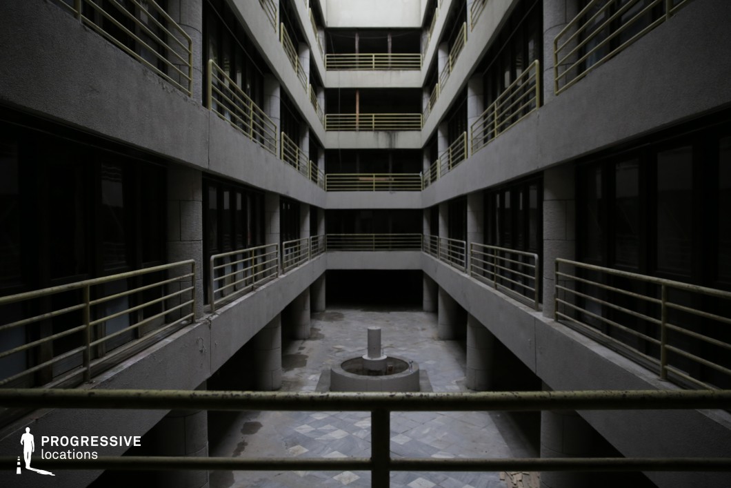 This image depicts an abandoned courtyard within a Brutalist-style residential complex. The space is surrounded by gray concrete walls, with several levels of balconies overlooking a central area. The yellow railings, though weathered, add a subtle contrast to the otherwise neutral tones of the concrete and glass. At the center of the courtyard, a dilapidated circular feature, likely a fountain, adds a focal point to the otherwise empty and worn space. The overall atmosphere is stark, cold, and industrial, with a sense of neglect. The unlit, quiet nature of the environment makes it ideal for scenes requiring a deserted or decaying urban setting, emphasizing isolation or the passage of time. The space’s symmetrical lines and open design could also serve as a backdrop for reflective, moody narratives or moments of quiet introspection in film or photography.