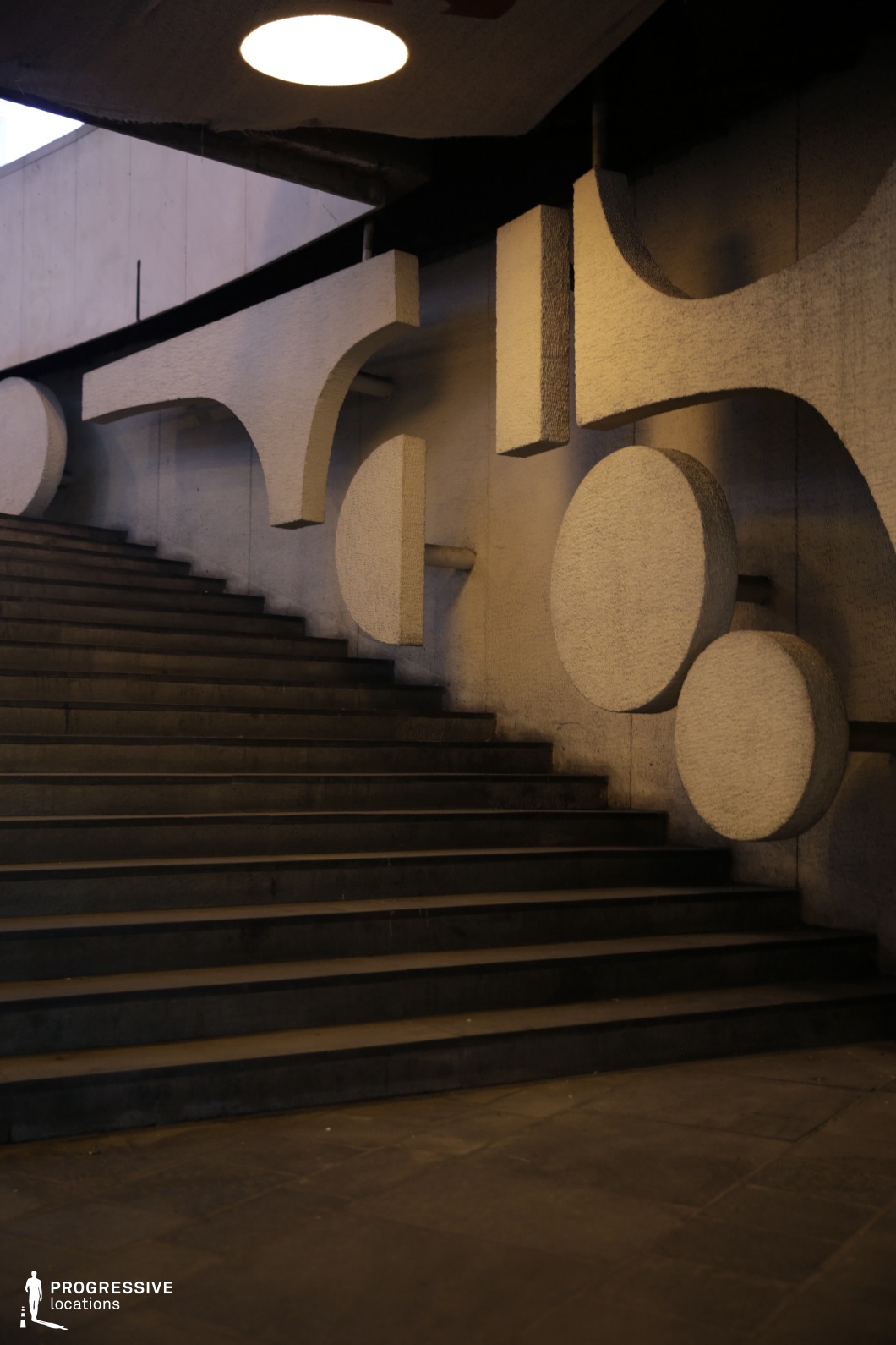 This image features a dramatic, sculptural staircase within a Brutalist architectural setting. The stairs are made of dark concrete, with bold, angular steps leading upwards. The most striking feature is the large, geometric concrete elements protruding from the wall beside the staircase. These abstract shapes, including circular and semi-circular forms, add a dynamic visual contrast to the solid, imposing stairs. Above, a circular light fixture illuminates the space, casting soft shadows that highlight the raw textures and sharp lines of the structure. The combination of the minimalist stairs and the striking sculptural forms creates a sense of depth and tension, ideal for cinematic scenes that require an industrial or modernist aesthetic. This location is perfect for atmospheric moments or visual storytelling where architecture plays a central role in setting the tone or highlighting the isolation and power of the space.