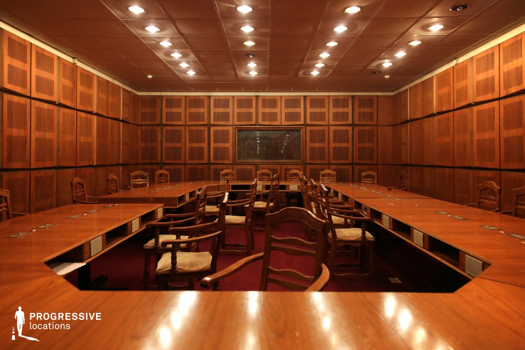 This image features a traditional conference room designed with rich wooden elements, evoking a sense of authority and formality. The room is furnished with a long wooden table surrounded by chairs, each with an intricately carved wooden frame and beige cushions. The walls are covered with wooden panels, each featuring a distinctive slatted pattern, adding a textural depth to the space. The warm wood tones are complemented by subtle, focused lighting from the ceiling, which highlights the polished surfaces of the table and walls. The atmosphere is both professional and timeless, making this space ideal for high-level meetings, discussions, or negotiations. The symmetrical arrangement and structured design of the room convey a sense of order, making it a fitting backdrop for scenes that require a setting of power, decision-making, or corporate authority. The overall design balances traditional elegance with modern functionality, offering a versatile setting for various creative and cinematic uses.