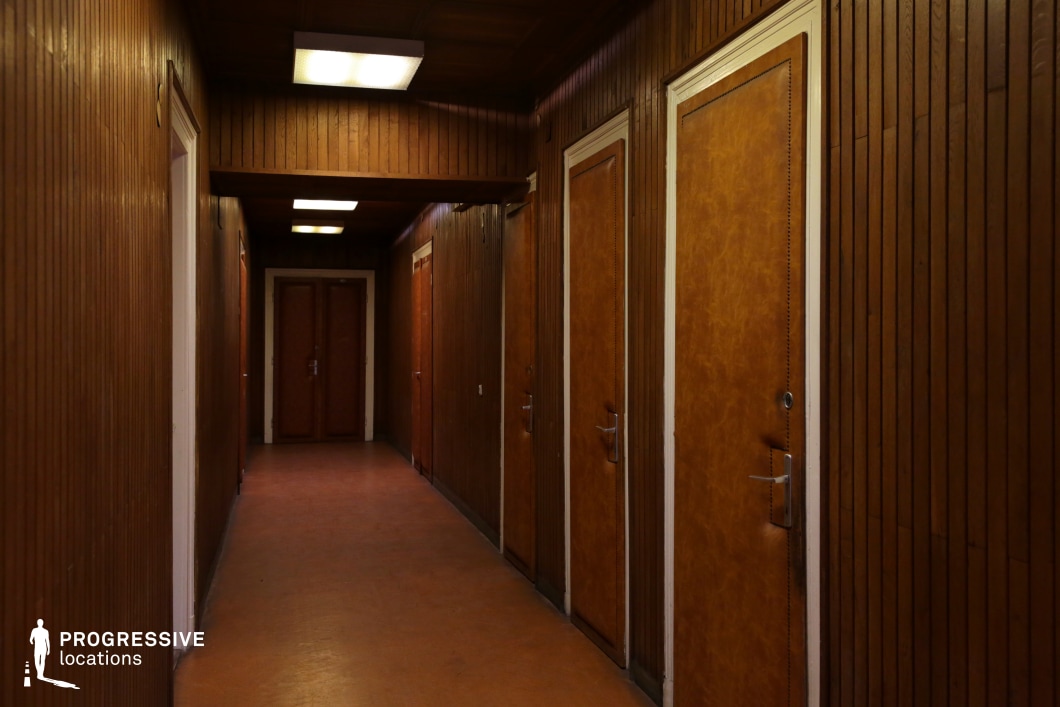This image features a narrow, retro-styled corridor with wooden paneling along the walls. The warm, dark wood is complemented by matching doors, each with simple, yet elegant handles and framed in white trim. The corridor is illuminated by fluorescent ceiling lights, casting a soft glow over the space, creating an atmosphere that feels both functional and slightly nostalgic. The repetition of the doors and the linear design of the space suggest a utilitarian, institutional environment, perhaps a hotel or an office building. The aged wooden finish and the uniformity of the doors contribute to a sense of time, evoking a mid-century modern style. This corridor would work well for scenes requiring a retro or industrial aesthetic, highlighting the passage of time or depicting mundane, everyday spaces with a sense of character and history. Its symmetry and simplicity make it an ideal backdrop for a variety of creative projects, from period dramas to urban-themed films.