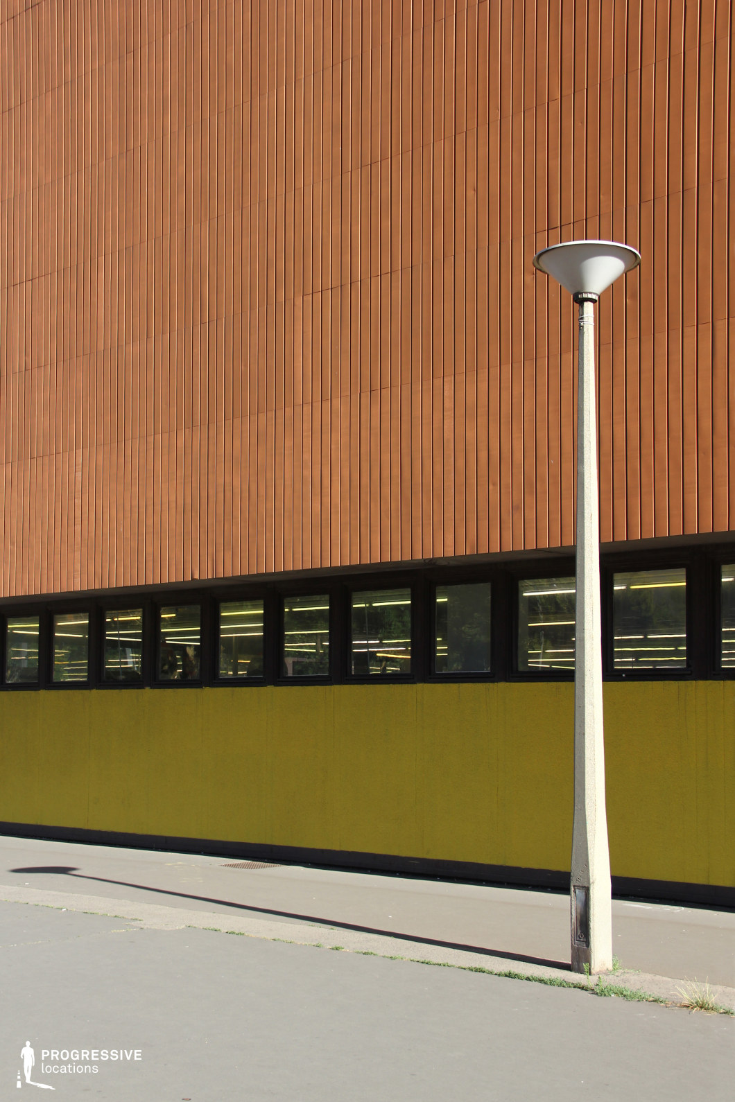 This image focuses on a section of a building's facade, highlighting the striking combination of textures and colors. The upper part of the building is adorned with vertical wooden panels that give the structure a sleek, modernist appearance. Below, a bold yellow section contrasts sharply with the earthy tones above, adding a layer of vibrancy to the design. The building's windows are set in a series of black frames, their uniform placement creating a rhythm across the facade. In the foreground, a tall streetlight stands at an angle, casting a long shadow on the pavement, which complements the clean, geometric lines of the structure. This location’s architectural style is ideal for scenes requiring an urban, mid-century modern aesthetic, making it suitable for commercial, industrial, or residential backdrops. The simplicity of the design and the interplay of colors and shapes offer a visually appealing environment for contemporary settings in film or photography.