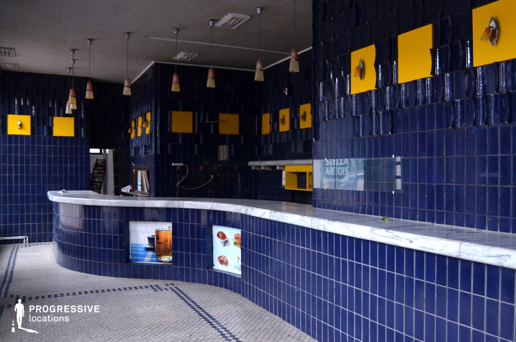 This image features a retro-style bar area, defined by vibrant blue ceramic tiles that cover both the walls and the counter. The tiles have a glossy finish, giving the space a polished yet vintage look. Interspersed with the deep blue tiles are bright yellow panels, adding a playful contrast and enhancing the bold, mid-century aesthetic. The bar itself is designed with a smooth marble countertop, which contrasts beautifully with the surrounding tiled surfaces. Above, pendant lights with soft orange shades cast gentle illumination over the space, enhancing the nostalgic atmosphere. The floor is adorned with white hexagonal tiles, further contributing to the overall retro vibe. This bar area, with its striking color scheme and geometric design, is ideal for scenes that evoke a sense of vintage charm, particularly for locations requiring a lively, vibrant setting or for narratives set in retro-themed establishments.