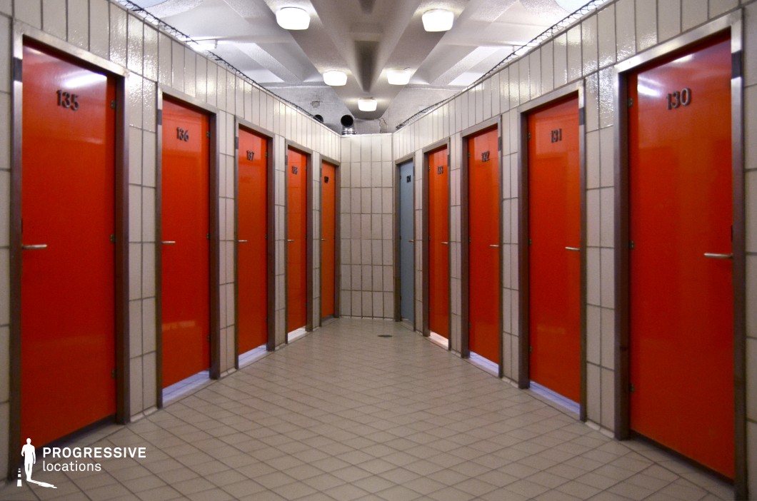 This image features a stark, geometric changing room with rows of vibrant orange doors, each numbered with black digits. The doors are framed by light-colored tiles that extend along the walls, creating a clean and functional aesthetic. The space is illuminated by ceiling lights, casting even light across the room and highlighting the uniformity of the layout. The repetitive pattern of the doors, arranged symmetrically along the corridor, gives the space a modern, almost industrial feel. The neutral tile floor complements the colorful doors, emphasizing the contrast between the bold orange and the subdued surroundings. This location, with its organized and straightforward design, would work well for scenes in institutional or recreational settings, such as gyms, spas, or locker rooms. The clean lines and vibrant accent color offer a visually appealing backdrop for moments of personal space or transitions in narrative.