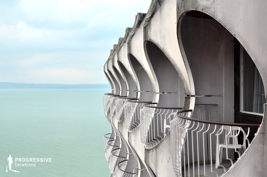 This image showcases a modernist architectural detail, featuring a series of concrete balconies with unique, circular cutouts, overlooking the tranquil expanse of a sea or lake. The design of the building highlights the geometric and industrial beauty of the architecture, with smooth white concrete and delicate metal railings forming a striking contrast against the vast, open water below. The repetition of the curved shapes along the facade creates a rhythmic pattern that adds a sense of dynamism to the otherwise minimalist structure. The serene water beyond enhances the calm, reflective mood of the setting, making it an ideal location for film scenes involving isolation, contemplation, or modernist environments. The combination of geometric design and natural surroundings also makes it a perfect backdrop for stories set in architectural-focused or scenic locations.